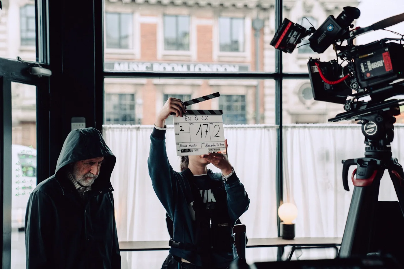 A crew member holds up a clapperboard in front of a hooded actor, with a cinema camera to the right