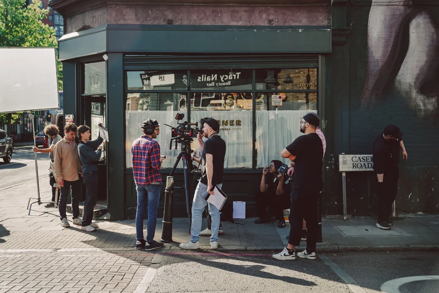 A film crew with a camera and reflector on the pavement outside a corner cafe with a mural on its wall