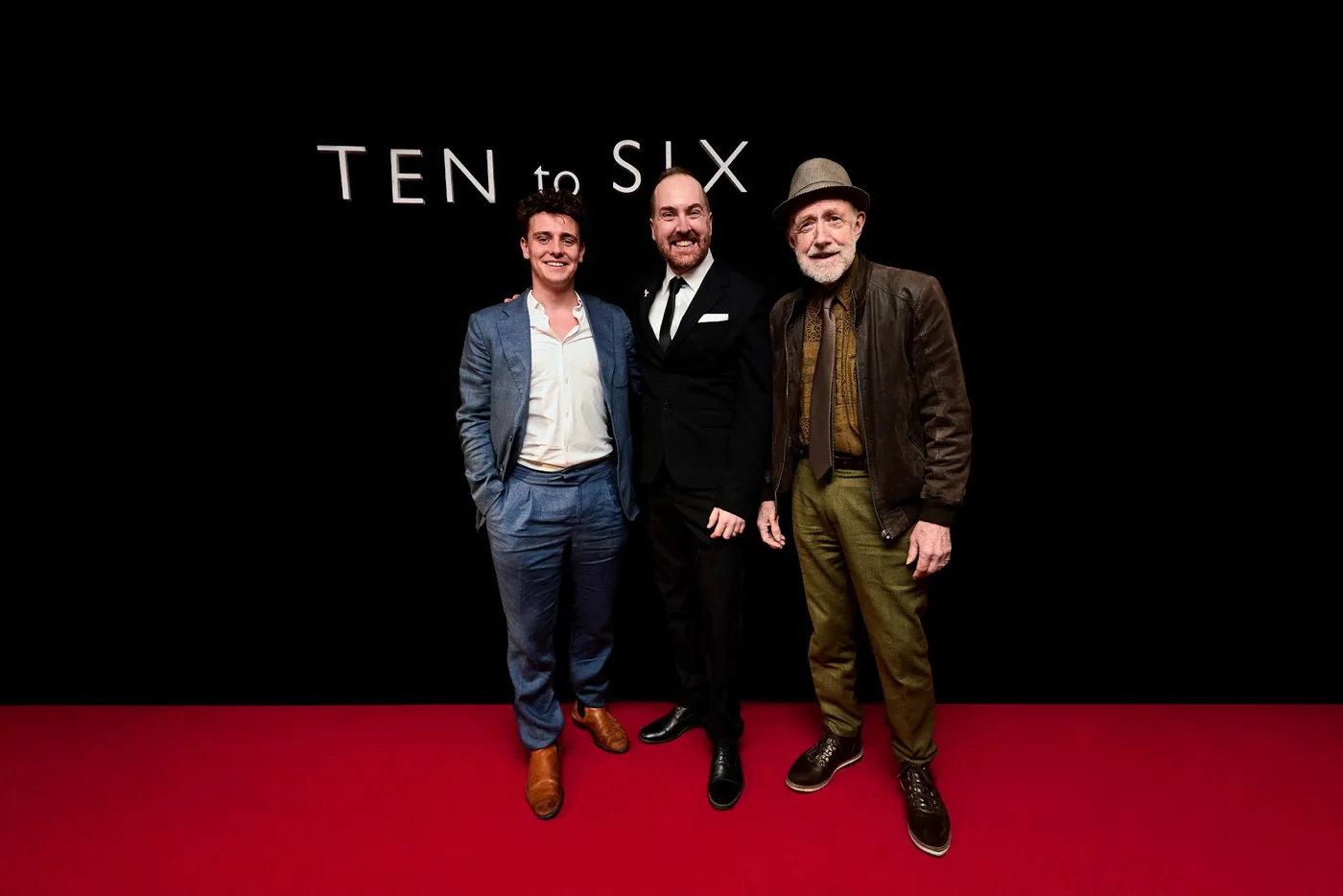 Three men in suits and a hat pose on a red carpet in front of a Ten to Six backdrop