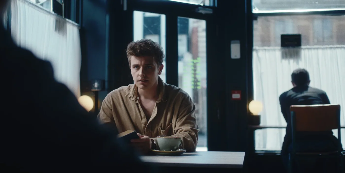 A young man in a beige shirt sits at a cafe table with a cup of coffee, in a still from Ten to Six