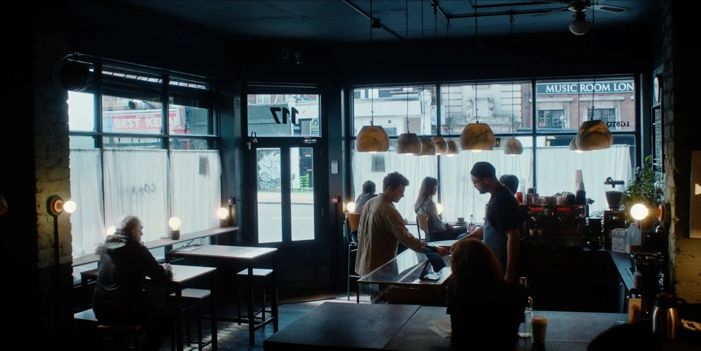 The interior of a cafe with large windows, hanging lamps and a few customers, in a still from Ten to Six