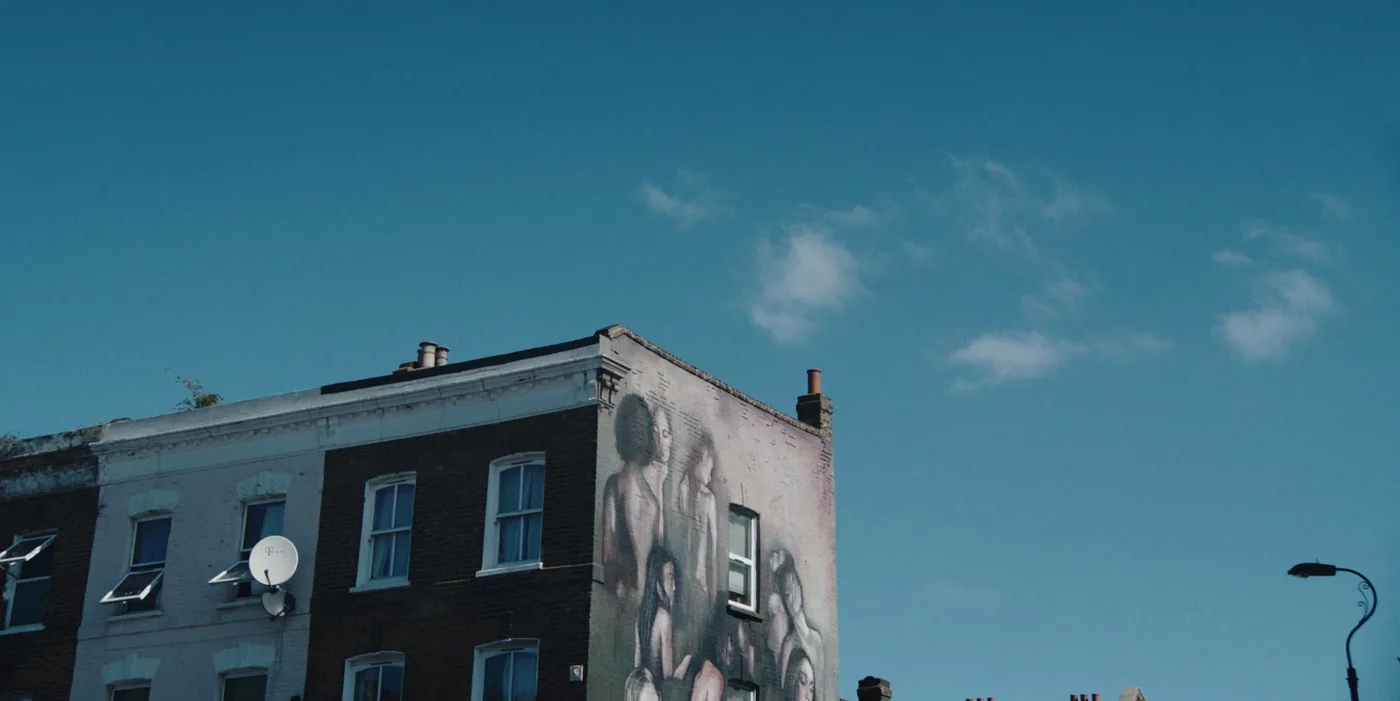 The top of a brick building with a large black and white mural on its side against a blue sky, in a still from Ten to Six