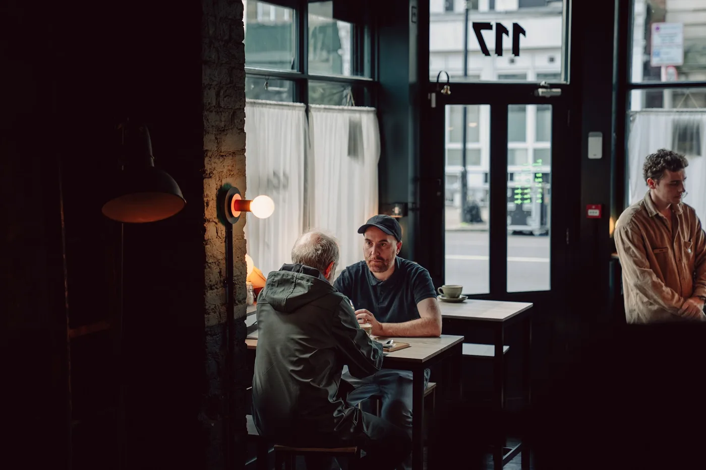 Actors at a table by the window of a cafe, with the number 117 on the glass