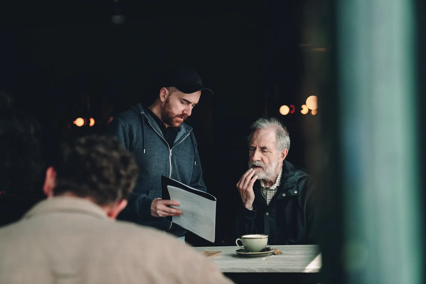 A man in a cap holding a tablet talks to an older actor seated at a cafe table with a cup of coffee