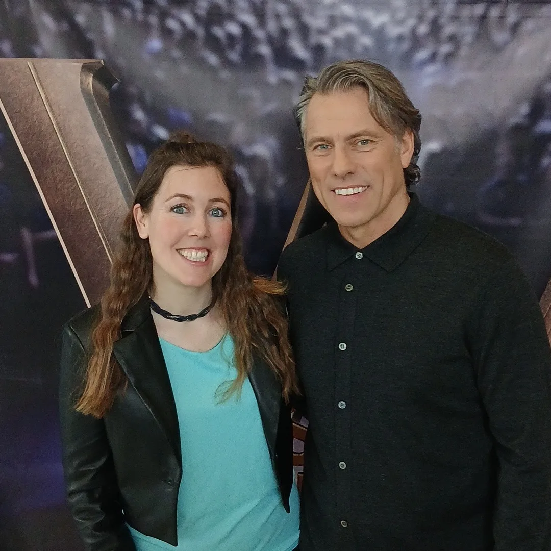 Kate Sweeney, in a black leather jacket and turquoise top, smiling beside John Bishop in a dark shirt, in front of a backdrop showing a large studio audience