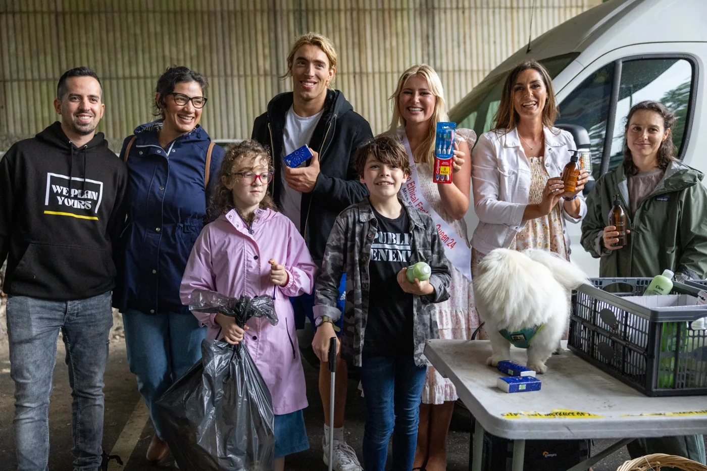 A group of adults and two children holding toiletries and a bin bag beside a table with crates and a white dog, in front of a white van