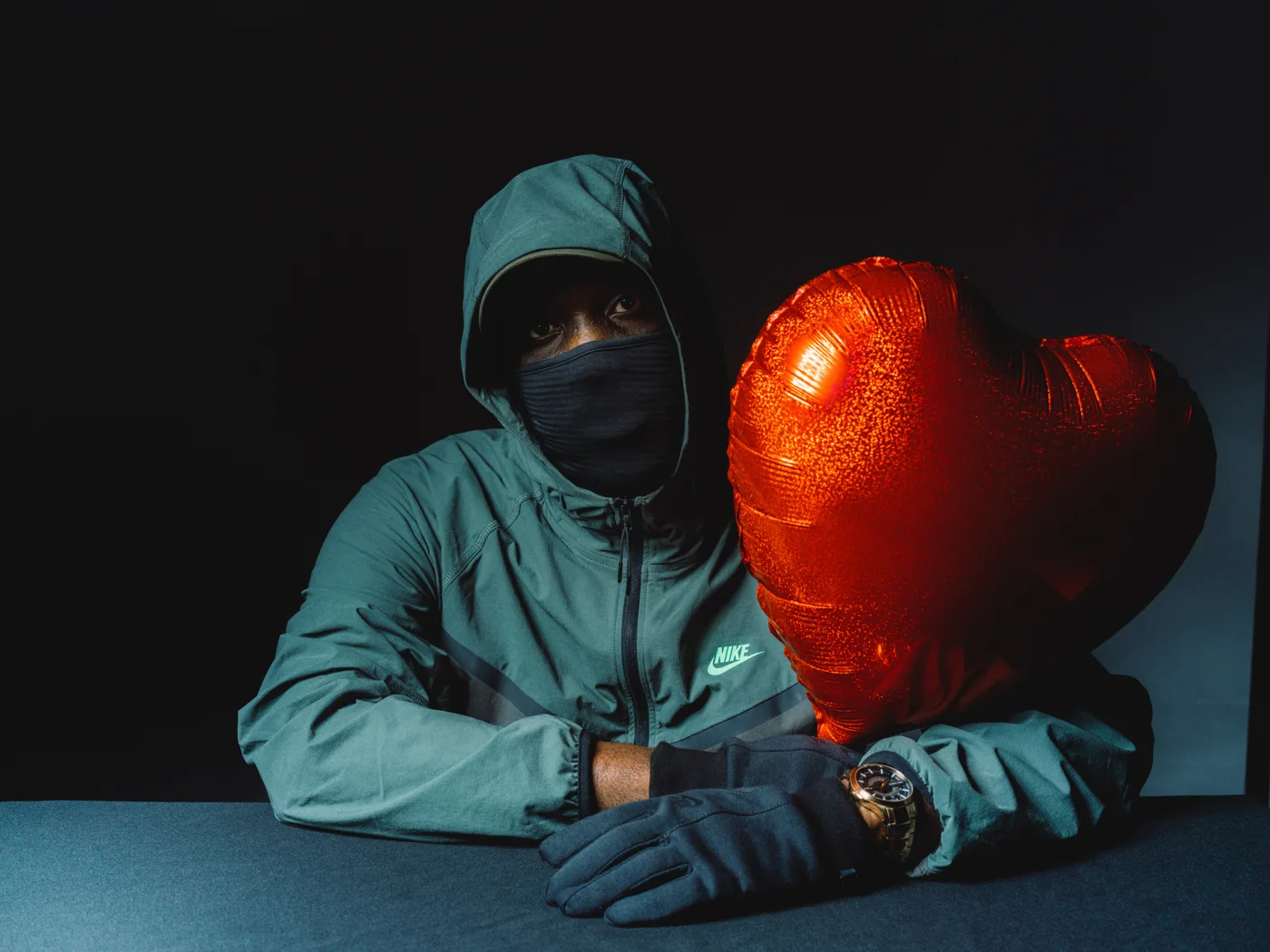 Triple OG Musiq in a green Nike tracksuit top, hood up and face covered, seated at a table with gloved hands and a red heart-shaped balloon beside him