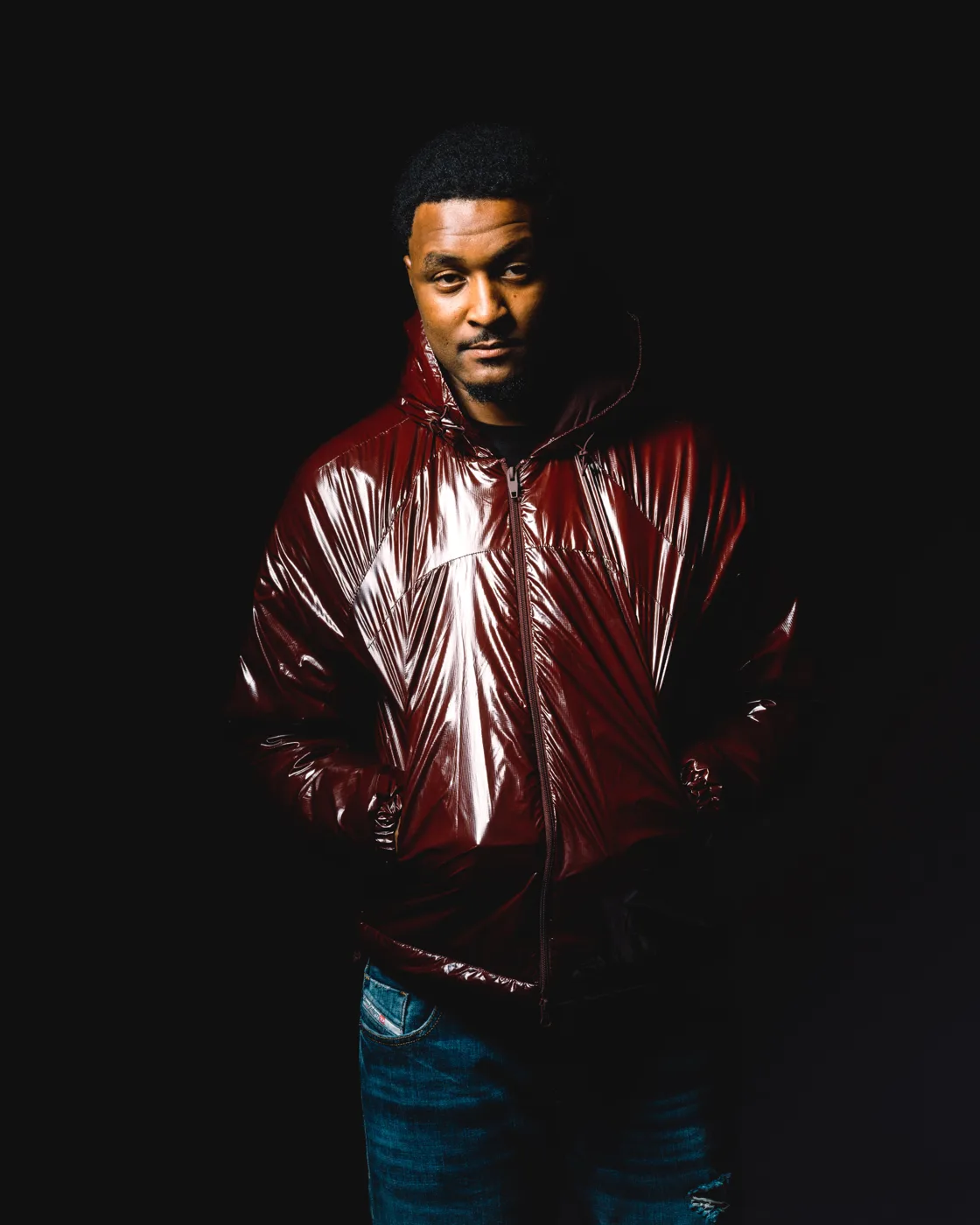 Scutla in a glossy maroon hooded jacket and jeans, standing against a black background