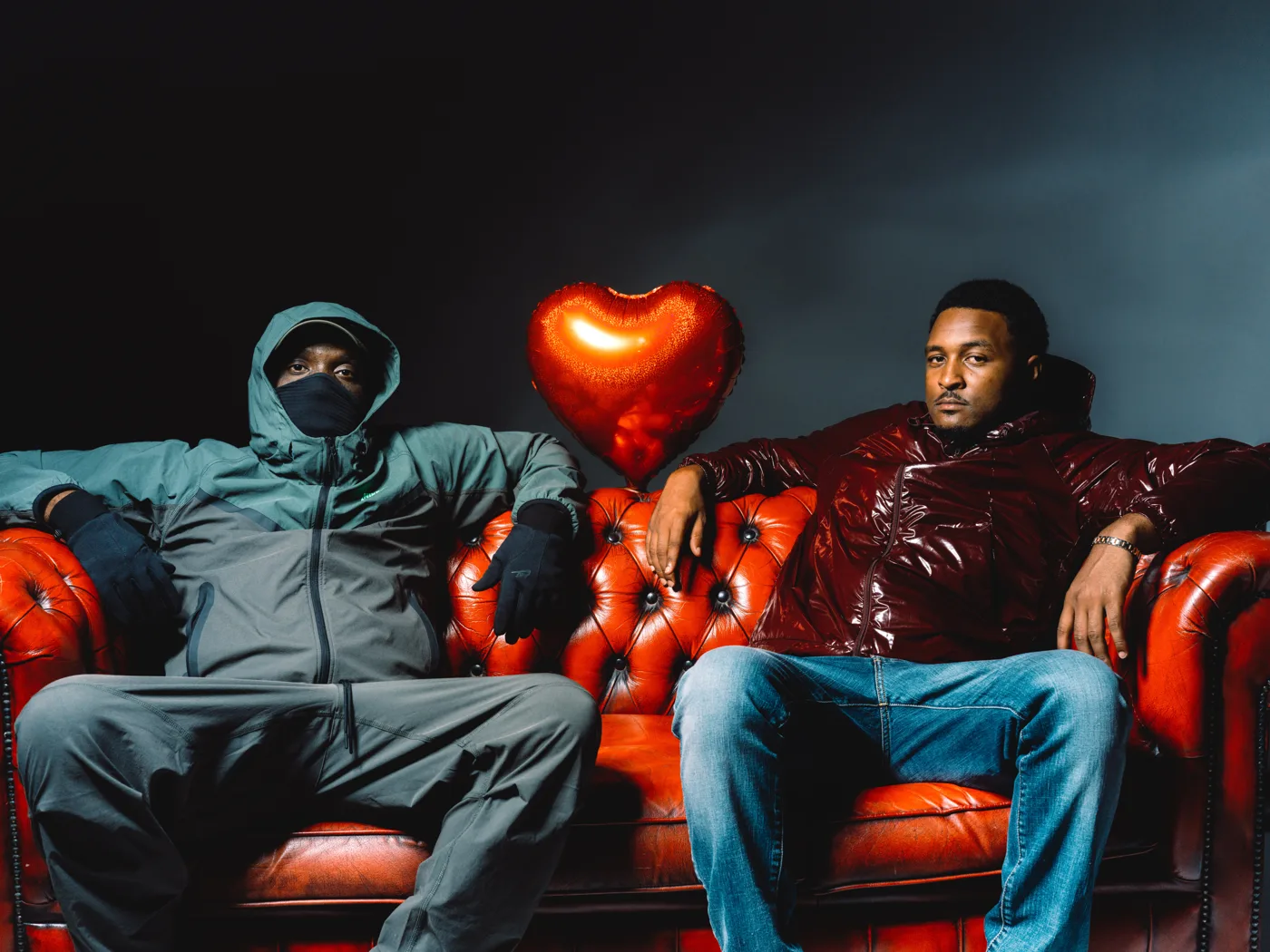 Triple OG Musiq, in the green tracksuit and face covering, and Scutla on a red leather sofa with a heart-shaped balloon